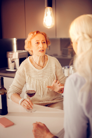 Alcoholic drink. Nice aged woman sitting with a glass of wine while talking to her friendの写真素材