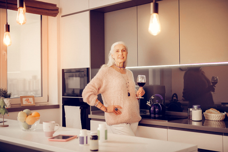At home. Joyful aged woman standing in the kitchen while holding a glass of wineの写真素材