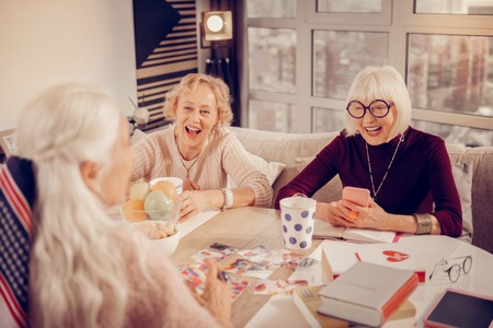 New technologies. Delighted aged woman looking at her smartphone while having a meeting with friends