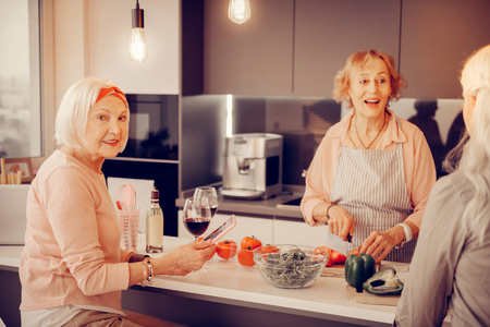 Internet addicted. Nice aged woman holding a tablet while sitting at the table in the kitchenの写真素材