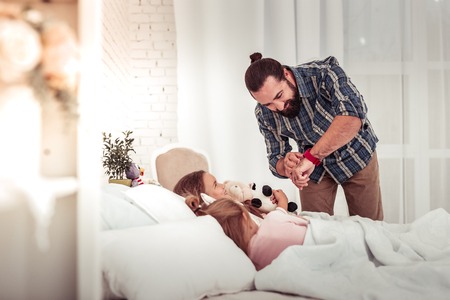 Time to sleep. Positive bearded man looking at the time while putting his daughters to sleepの写真素材