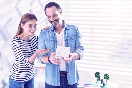 Fantastic design. Cheerful young woman standing next to the smiling man and pointing to the miniature house in his handsの写真素材