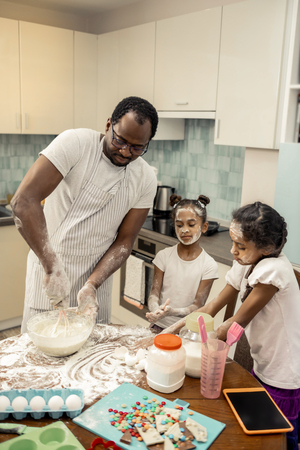 Cook pie together. Father wearing striped apron beating whites while cooking pie with his daughtersの写真素材