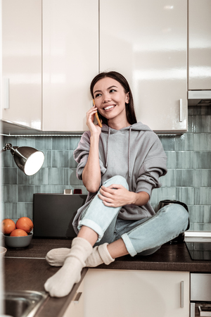 Speaking on phone. Slim appealing dark-haired woman sitting in the kitchen and speaking on the phoneの写真素材