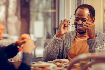 Cheerful pleasant man putting his glasses on sitting near window in restaurantの写真素材