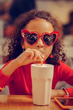 Funny cute girl wearing red sunglasses drinking milkshake in cafeteriaの写真素材