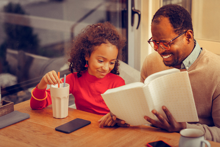 Father laughing. Cheerful father wearing glasses laughing while reading book for daughterの写真素材