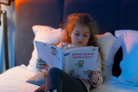 Before sleep. Small cute long-haired girl in a sweater looking interested while reading a book in bedの写真素材