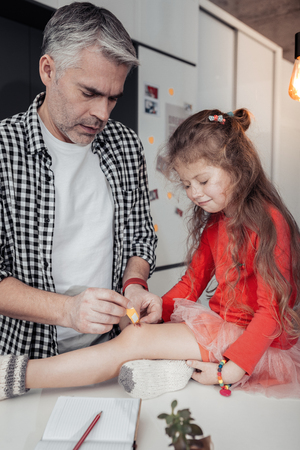 Girl on the table. Father wearing a red watch fixing a patch on the wounded kneeの写真素材