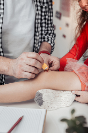 Crying small girl. Tall grey-haired father wearing a red watch on his hand curing his daughters kneeの写真素材