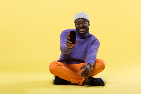 Life enjoyment. Pleased model sitting over yellow background and holding telephone in right handの写真素材