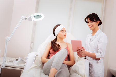 Professional cosmetology. Delighted positive cosmetologist standing near her client while looking at the tablet screenの写真素材