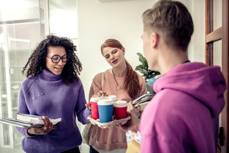 Coffee time. Two beautiful stylish students feeling pleased while taking coffee from their friendの写真素材