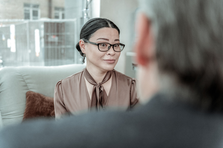 Surprised. Dark-haired elegant woman wearing a brown satin blouse looking surprised while speaking in the office with her partnerの写真素材