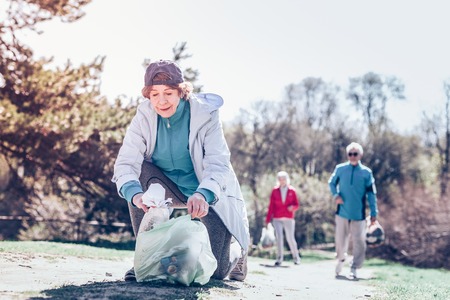 Park clean. Pleasant dark-haired retired woman wearing cap while cleaning the park at the weekendの写真素材