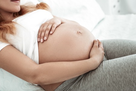 Love and tenderness. Caring long-haired woman covering her flawless round bronzed stomach with both tidy handsの写真素材