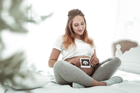 Child inside. Pleasant happy future mother observing photo result of ultrasound procedure and attaching it to round bellyの写真素材