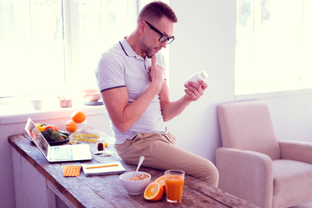 Man hesitating. Bearded mature man wearing glasses hesitating about drinking food supplements while reading ingredientsの写真素材