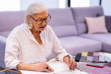 Working boss. Comely smiling elderly left-handed woman in glasses and beautiful fancy earrings making designer sketch in sitting at the office desk.の写真素材