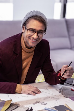 Adult working man. Smiling fancy adult gentleman in black-rubbed glasses, knitted hat and modish jacket energetically working on drawings in officeの写真素材
