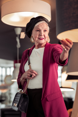 Fancy elderly lady portrait. The portrait of elegant sophisticated woman in advanced years in black leather beret, massive jewelry and cross-body black bag checking the lamp quality in storeの写真素材
