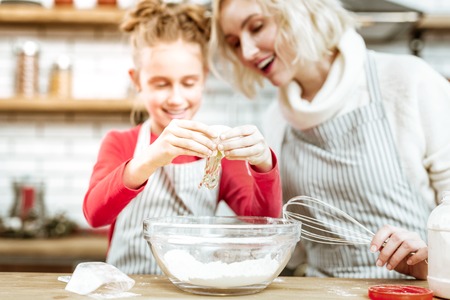 Cooking with mother. Happy good-looking mother attentively looking on hardworking daughter while she putting egg in bowlの写真素材