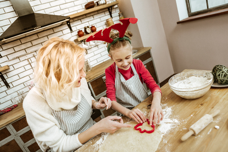 Using baking tools. Smiling positive little child in striped apron learning cooking aspects with her supportive motherの写真素材