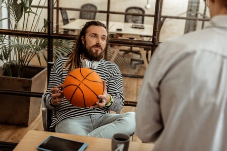 Favourite game. Delighted nice man holding a basketball ball while sitting in the chairの写真素材