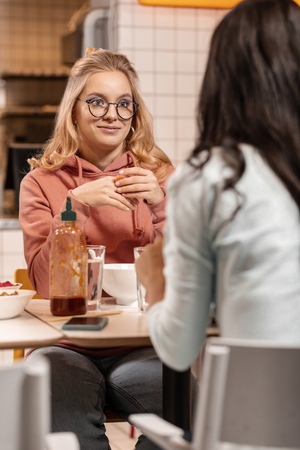 Pleasant time. Attractive confused girl in glasses listening to her female friend while having lunch.の写真素材