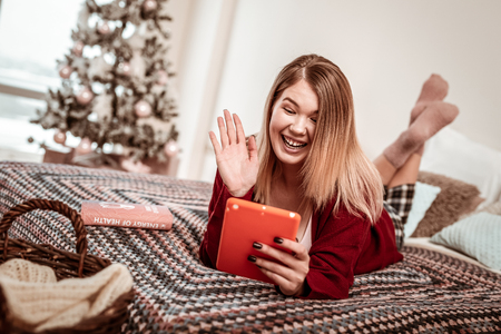 Having online call. Coquettish young girl using tablet for video connection while lying in bedの写真素材