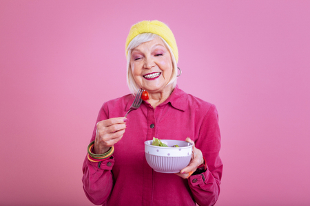 Cheerful lady. Cheerful elderly lady wearing pink dress feeling positive while eating healthy saladの写真素材