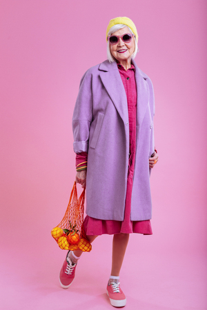 Bright sunglasses. Stylish retired woman wearing bright sunglasses holding string bag with fruitsの写真素材