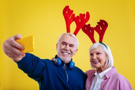Photo with wife. Retired man holding yellow phone making photo with his wife wearing cute deer racksの写真素材