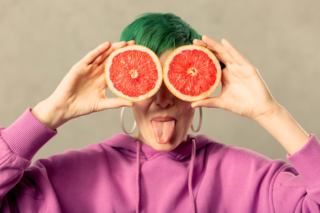So playful. Nice happy young woman showing her tongue while standing with grapefruit halvesの写真素材