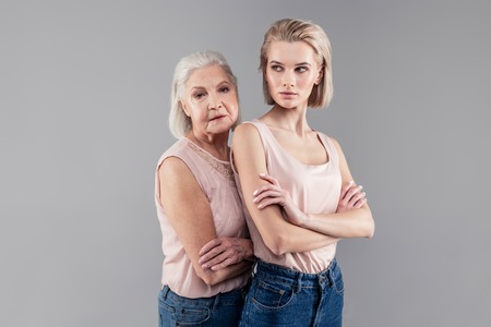 Serious faces. Senior grey-haired mother leaning on her daughter back while standing behindの写真素材