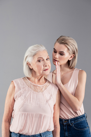 Attentively listening. Accurate serious blonde girl carefully whispering information to her curious grey-haired old motherの写真素材