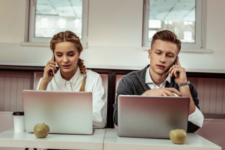 Cheerful young guys. Focused good-looking colleagues sitting close to each other and answering on phone callsの写真素材