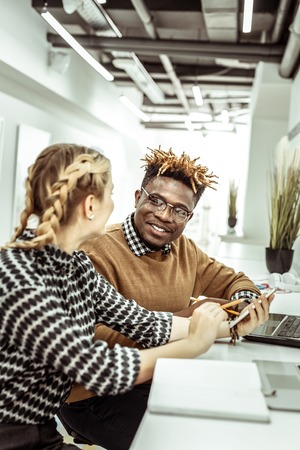 Man pleasantly looking. Good-looking woman with two braids pointing on smartphone screen while informing her curious friendの写真素材