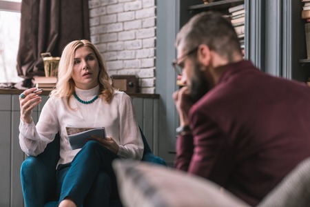 Psychologist in necklace. Blonde-haired psychologist wearing white blouse and blue necklace listening to her clientの写真素材