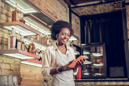 Afro-American bartender with tablet. Charming bewitching curly-haired beaming Afro-American bar employee in stylish green apron filling in order form with red-colored tabletの写真素材