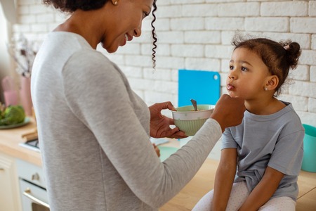 Be slowly. Cute curly-haired woman standing in semi position and feeding her daughterの写真素材