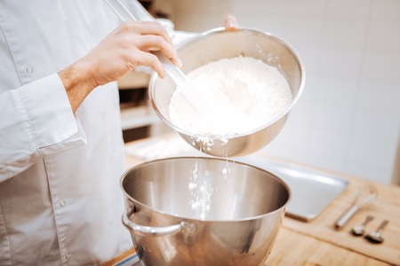 Cooking bread. Close up of professional young baker in uniform adding some flour while cooking bread