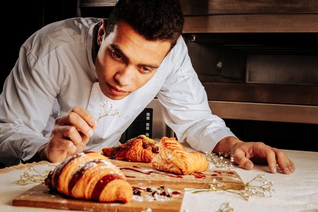 Looking at desserts. Young dark-haired baker leaning on table and looking at amazing dessertsの写真素材