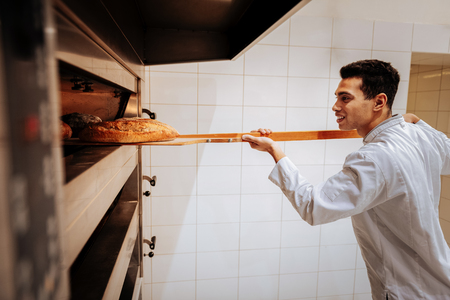 Taking baguettes. Dark-haired talented baker smiling while taking baguettes out of oven