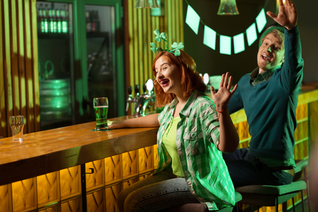 Meeting friends. Fair-haired young handsome man in a green hat and a red-haired girl with shamrock symbols on her head waving to their friendの写真素材
