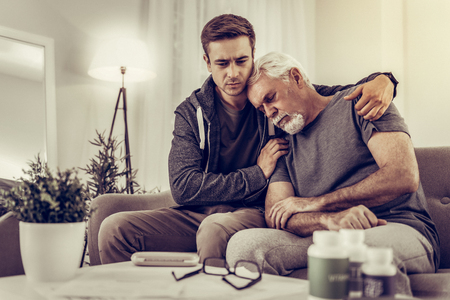 Support for sick person. Heartsore adult grieving grandchild in grey hoodie supportingly hugging and comforting his elderly silver-haired granddad after finding out his diseaseの写真素材