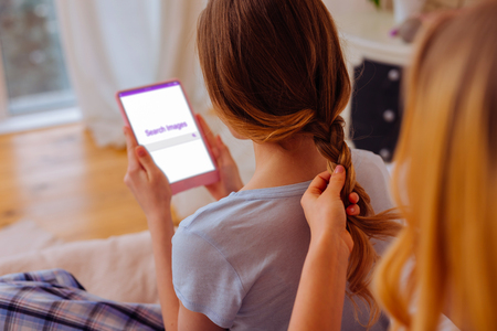 Making braid. Loving older sister making braid for her teenage sibling using tablet and the Internetの写真素材