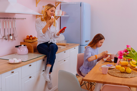 Sitting near cooker. Older blonde-haired sister sitting near the cooker and reading online news during breakfastの写真素材