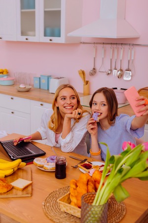 Selfie time. Teenage girl holding pink smartphone while making selfie with older sister sitting with laptopの写真素材