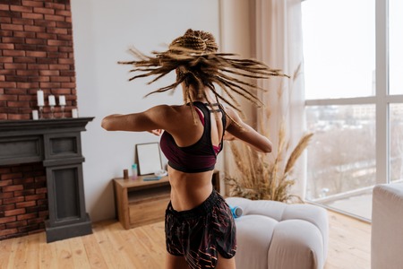 Stretching back. Active woman with nice little braids stretching her back at home near the window